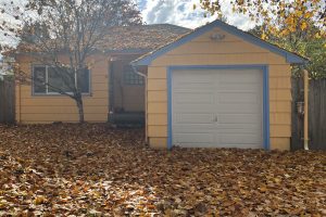 A small yellow house and detached garage surrounded by fallen autumn leaves covering the entire front yard and driveway.