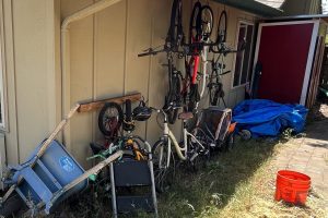 Outdoor clutter including bikes, tools, and yard equipment stored directly against the siding of a house.